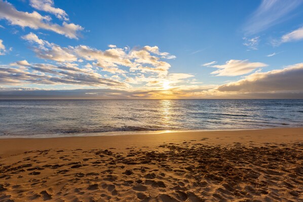 Una playa con huellas que conducen al agua, un cielo despejado y el sol poniéndose en el horizonte.