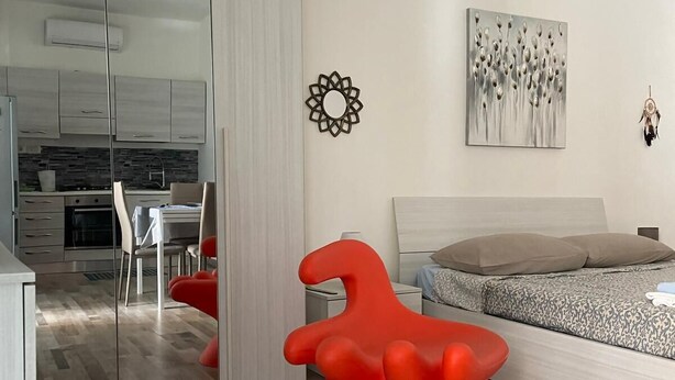 Ein modernes Schlafzimmer mit einem Bett, einem roten Sessel, einem Wandbild und einer Küche im Hintergrund.
