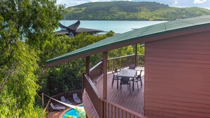 A wooden deck with a pool, lounge chairs, and a table overlooking a body of water and hills. - Casuarina Cove 6 (Hamilton Island)