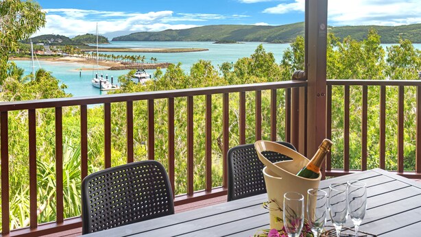 A balcony with a table set for a meal, a bottle of champagne, and glasses, overlooking a bay with boats and greenery. - Casuarina Cove 6 (Hamilton Island)