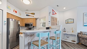 A compact kitchen with a refrigerator, microwave, and wooden cabinets. There are two blue chairs with woven seats. - Beacon Waves - Beach condo w/ Pool (Kill Devil Hills)
