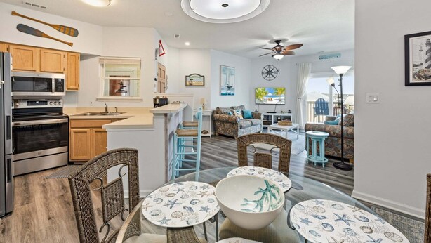 A compact kitchen with modern appliances, a dining area with wicker chairs and a round table, and a living room with a sofa and a TV. - Beacon Waves - Beach condo w/ Pool (Kill Devil Hills)