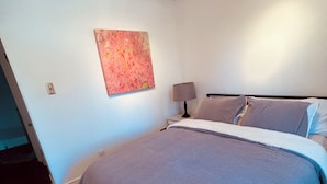 Quarto com cama, mesa de cabeceira, luminária, pintura e tapete estampado.