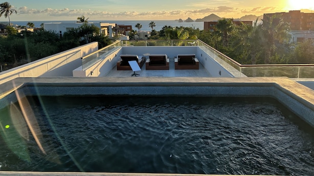 En pool på taket med solstolar, ett glasräcke och utsikt över havet och byggnaderna.