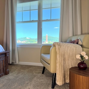 A room with a large window, a beige armchair, a small wooden side table with a vase, and a dark wooden desk. - Overlook at Liberty: Sleeps 10, <1 Mile to LU + Campus & Mountain Views (Lynchburg)