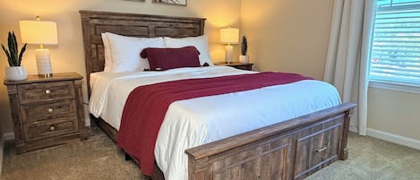 Un dormitorio con una cama de madera, dos mesitas de noche, una mesita auxiliar, un ventanal con persianas y dos lámparas fijadas a la pared.