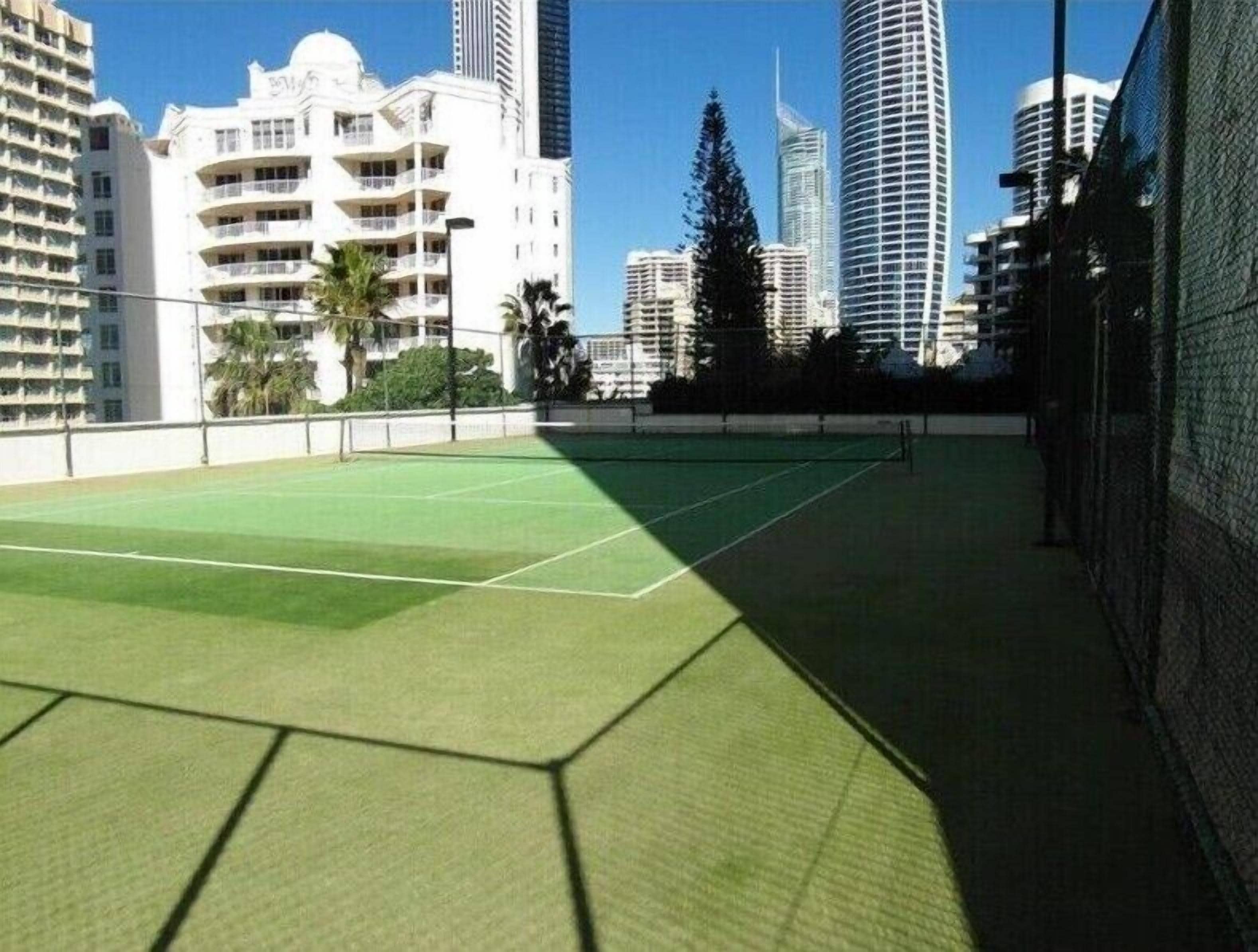 An indoor tennis court with a green surface, surrounded by high-rise buildings.