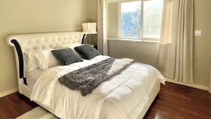 Une chambre à coucher avec un grand lit, une table de chevet, une lampe, une fenêtre avec des rideaux et un plancher en bois.