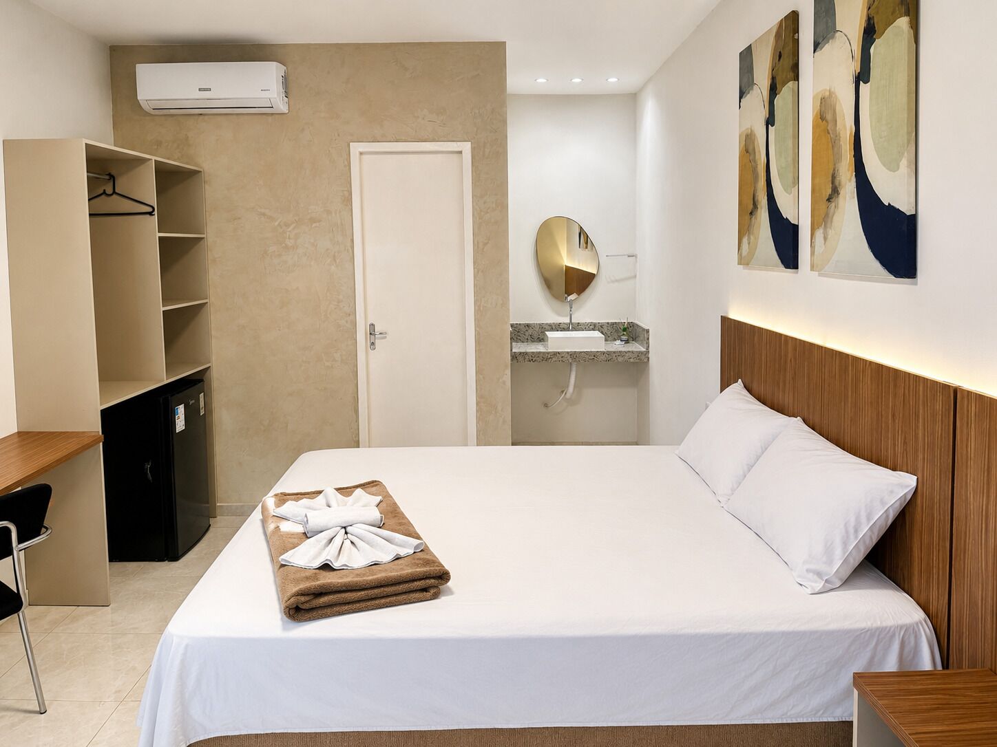 Ett hotellrum med en säng, ett skrivbord, en garderob och ett badrum med en spegel, samt abstrakt konst på väggarna.