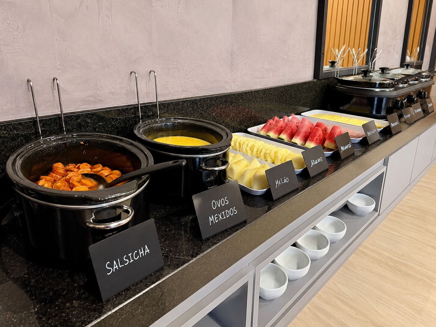 Un buffet proposant différents plats, chacun étant clairement identifié, notamment des « salsichas » et du « melão ».