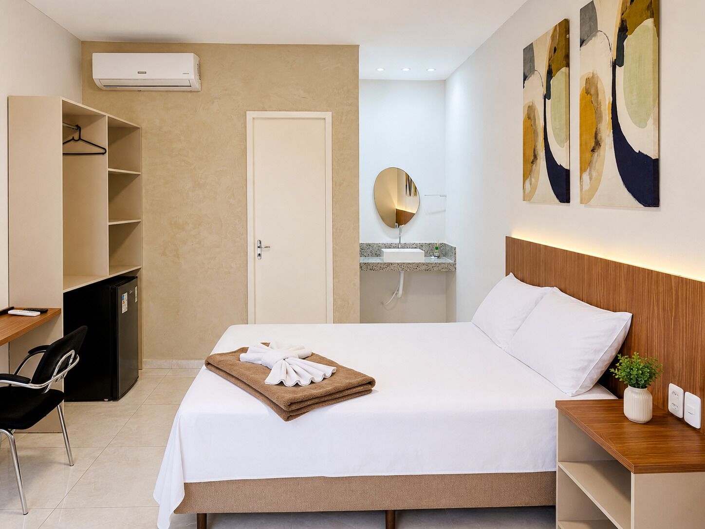 A hotel room with a bed, a desk, a chair, a wardrobe, a mirror, and a sink.