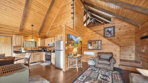 A log cabin interior with a kitchen, dining area, and living room, featuring wooden walls, a high ceiling, and a fireplace. - Family-Friendly Blowing Rock Retreat | Fire Pit, Game Room, & Cozy Mountain Views (Lenoir)