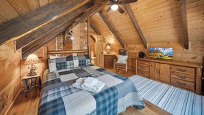 A cozy wooden cabin bedroom with a bed, a desk, a chair, a television, and a ceiling fan. - Family-Friendly Blowing Rock Retreat | Fire Pit, Game Room, & Cozy Mountain Views (Lenoir)