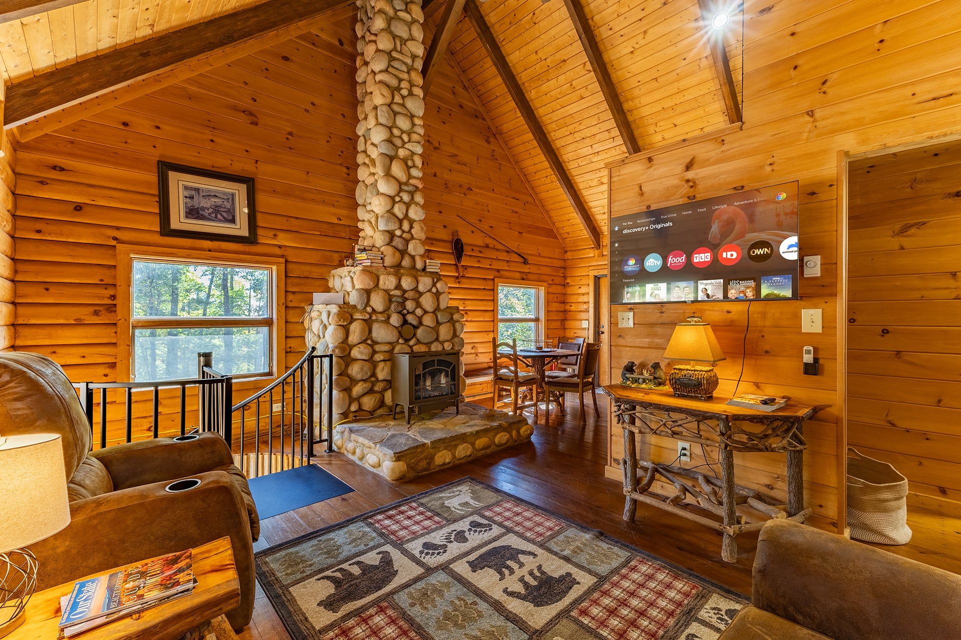 A log cabin interior with a stone fireplace, wooden walls, a dining area, and a television mounted on the wall.