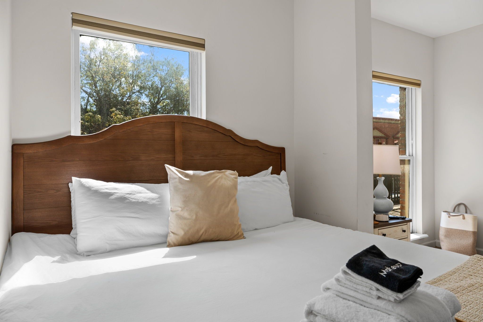 A neatly made bed with white and beige pillows, a wooden headboard, and a window with a view of trees.