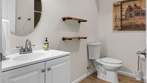 Un baño con un lavamanos blanco, un inodoro y un espejo. Hay estantes de madera sobre el inodoro y un cuadro en la pared.