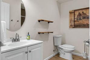 A bathroom with a white sink, a toilet, and a mirror. There are wooden shelves above the toilet and a painting on the wall.
