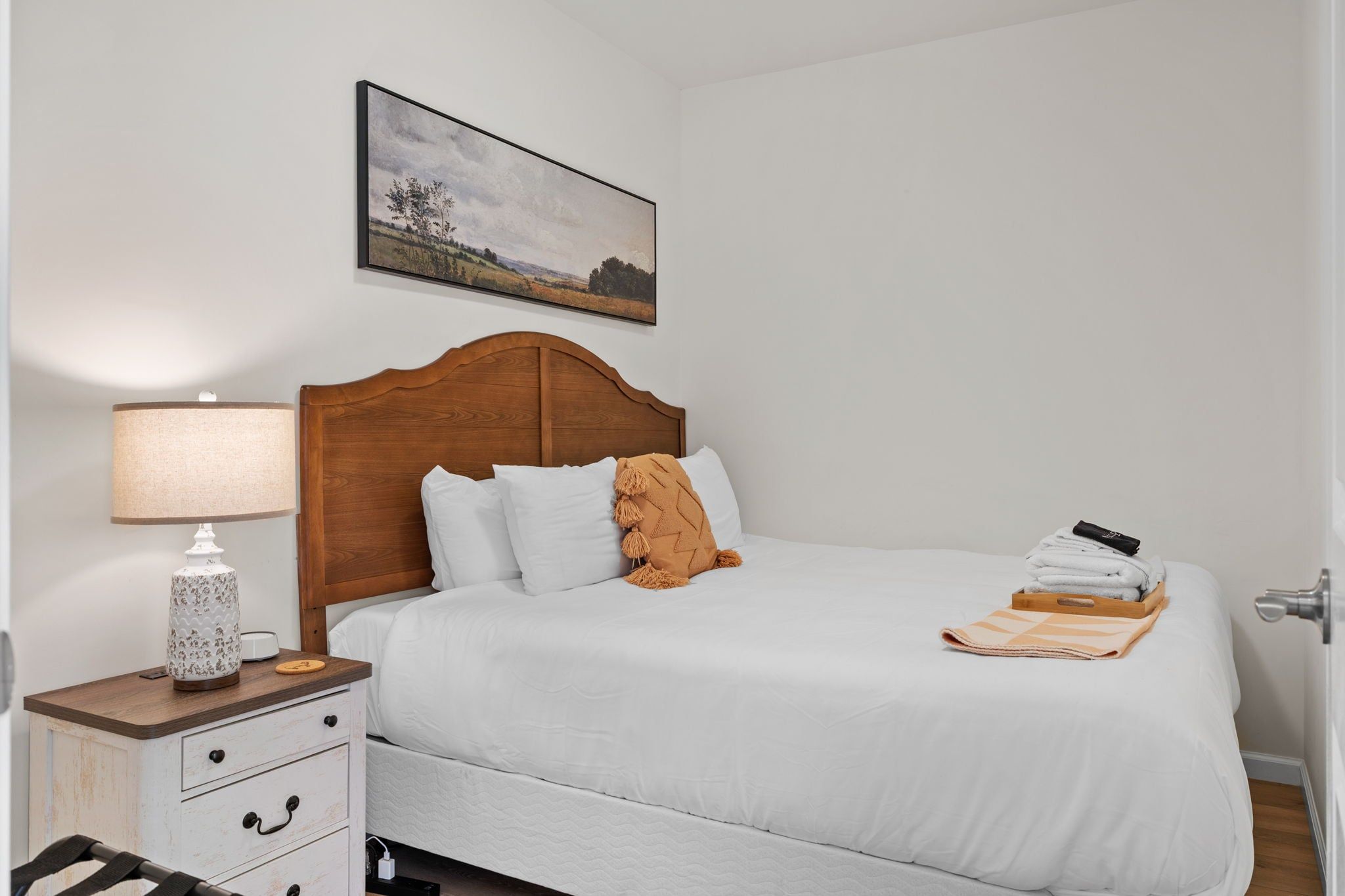 Une chambre à coucher avec une tête de lit en bois, du linge de lit blanc, une lampe de chevet, une petite table ornée d’un objet décoratif et un tableau représentant un paysage, encadré, accroché au mur.