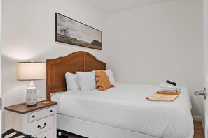 A bedroom with a wooden headboard, white bedding, a bedside lamp, a small table with a decorative item, and a framed landscape painting on the wall. - Hearth & Harvest | Historic St. Charles 2BR (Saint Charles)