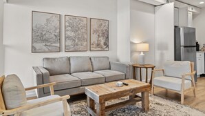 Una sala de estar moderna con un sofá gris, una mesa de centro de madera y una mesita auxiliar con una lámpara. Se ve una cocina con un refrigerador y armarios al fondo.