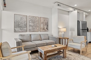 A modern living room with a grey sofa, wooden coffee table, and a small side table with a lamp. There is a kitchen area with a refrigerator and cabinets in the background.