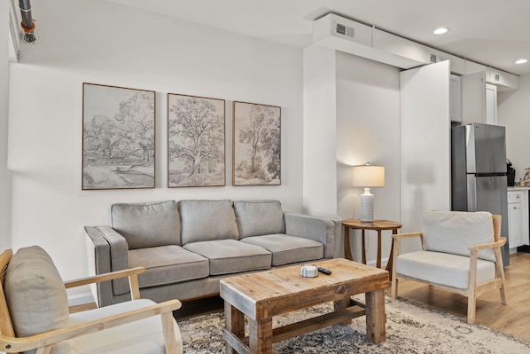 Una sala de estar moderna con un sofá gris, una mesa de centro de madera y una mesita auxiliar con una lámpara. Se ve una cocina con un refrigerador y armarios al fondo.