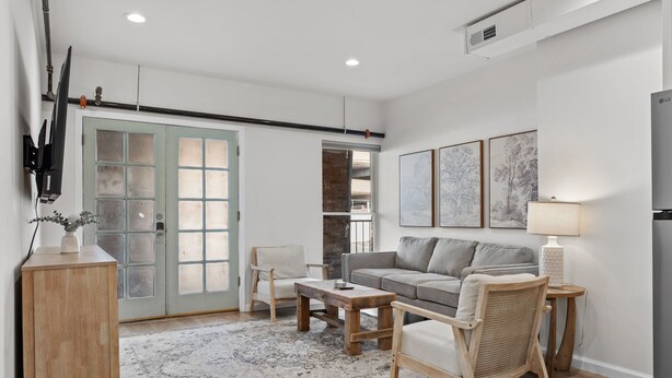 Una sala de estar moderna con un sofá, una mesa de centro y una silla de mimbre. Se observa un mueble de madera y un televisor montado en la pared.