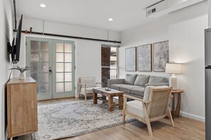A modern living room with a sofa, coffee table, and a wicker chair. There is a wooden cabinet and a TV mounted on the wall. - Hearth & Harvest | Historic St. Charles 2BR (Saint Charles)