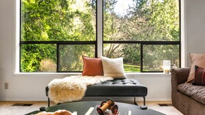 A cozy living room with a large window, a black sofa, a green chair, and a coffee table with books and a spool of twine.