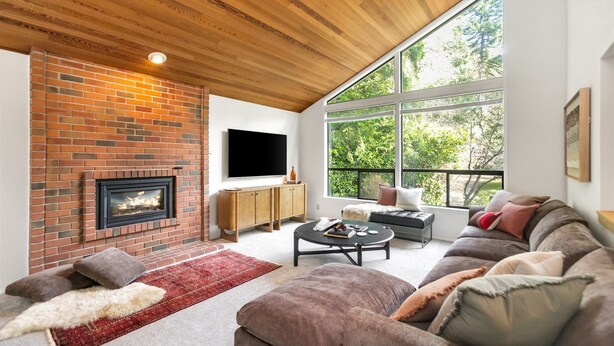 A living room with a brick fireplace, a large window, a sofa, and a television.