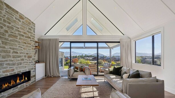 A spacious living room with a stone wall, a fireplace, a large sofa, a coffee table, and a view of mountains through large windows. - Miharo Mountain Haven | Mountain Views & Outdoor Fire (Wānaka)