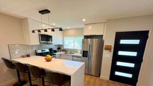 A modern kitchen with a central island, bar stools, stainless steel appliances, and a black-framed door. - 3-Bed, 4-Bath Modern Home - Open Concept, Fully Remodeled & Brand-New Furnished (Los Angeles)