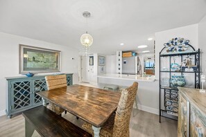 Uma sala de jantar com mesa de madeira e cadeiras de vime, um candelabro e uma cozinha ao fundo.