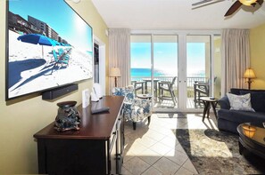 Ein Zimmer mit Fernseher, der eine Strandansicht zeigt, einem Balkon mit Meerblick, einer Couch und einem kleinen Tisch.