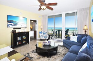A living room with a television, a ceiling fan, a sofa, and a view of the ocean through large windows. - NEW  Waters Edge 310 • Beautiful Beach Condo, Premium Views, Covered Pkg, Free Beach Service (Fort Walton Beach)