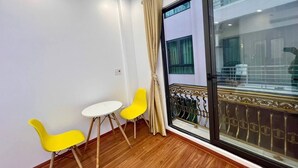 Ein Zimmer mit Holzboden, einem weißen Tisch, zwei gelben Stühlen, einem beige farbenen Vorhang und einer Glastür zum Außenbalkon.