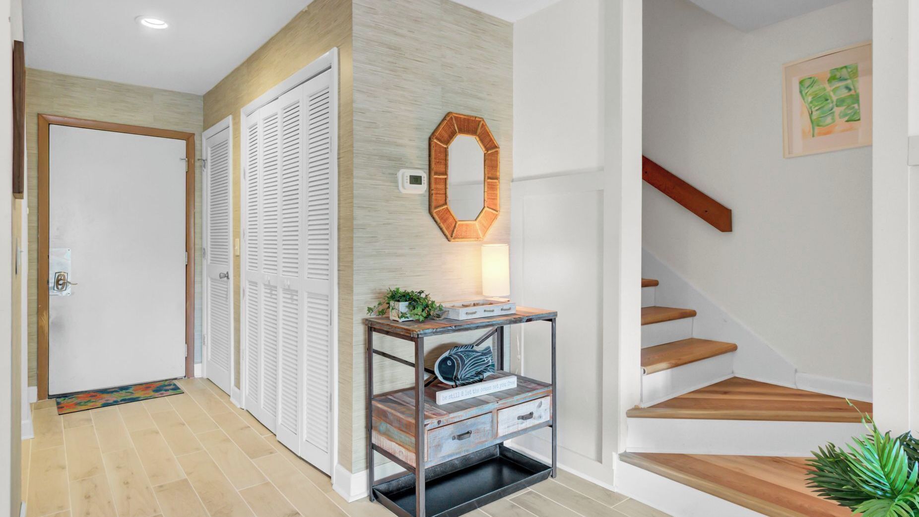 Un pasillo con piso de madera, puertas blancas, un espejo montado en la pared, una mesita con lámpara y una escalera de madera.