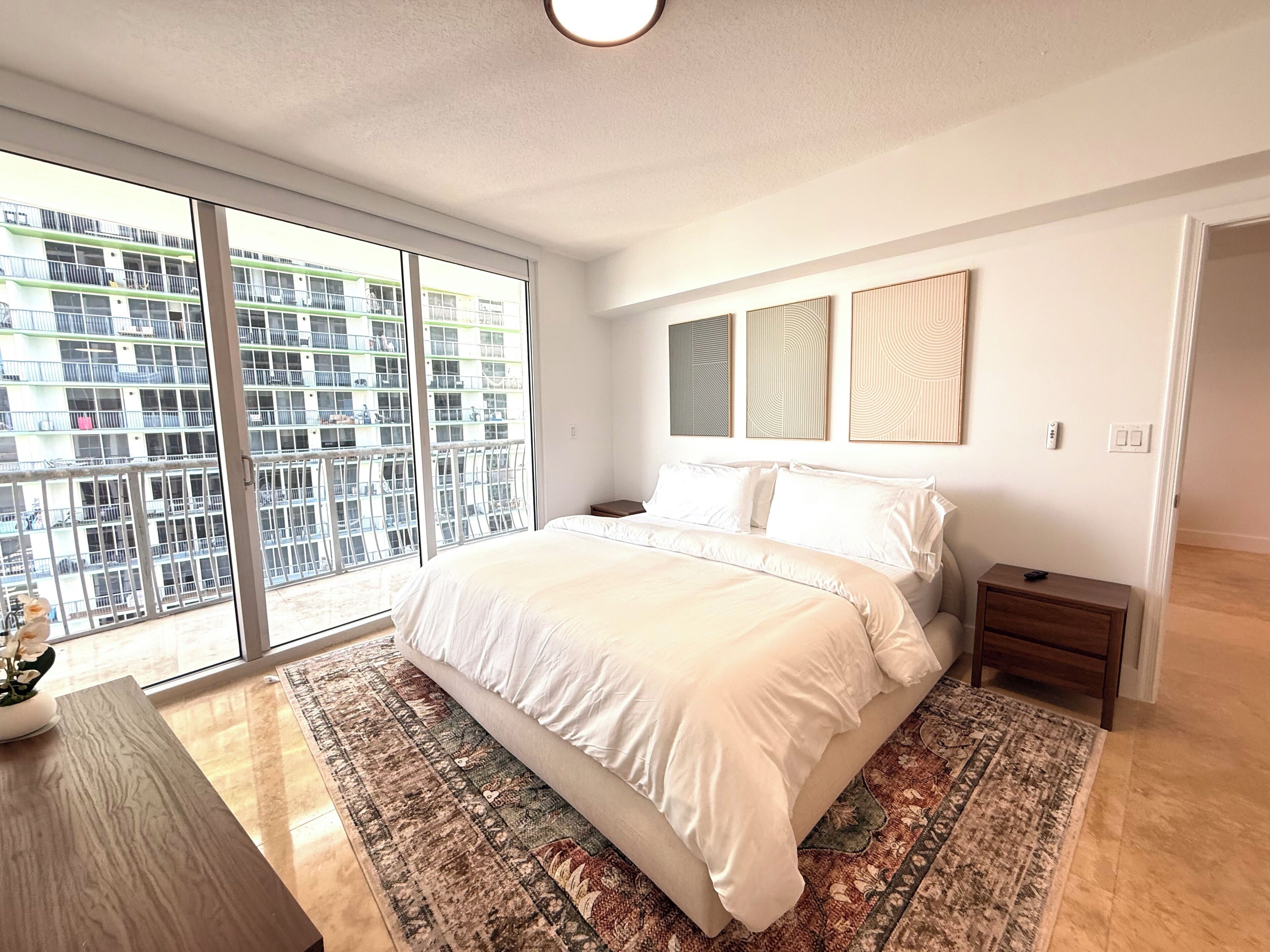 A modern bedroom with a large bed, a nightstand, a wooden side table, and a view of a building through floor-to-ceiling windows.