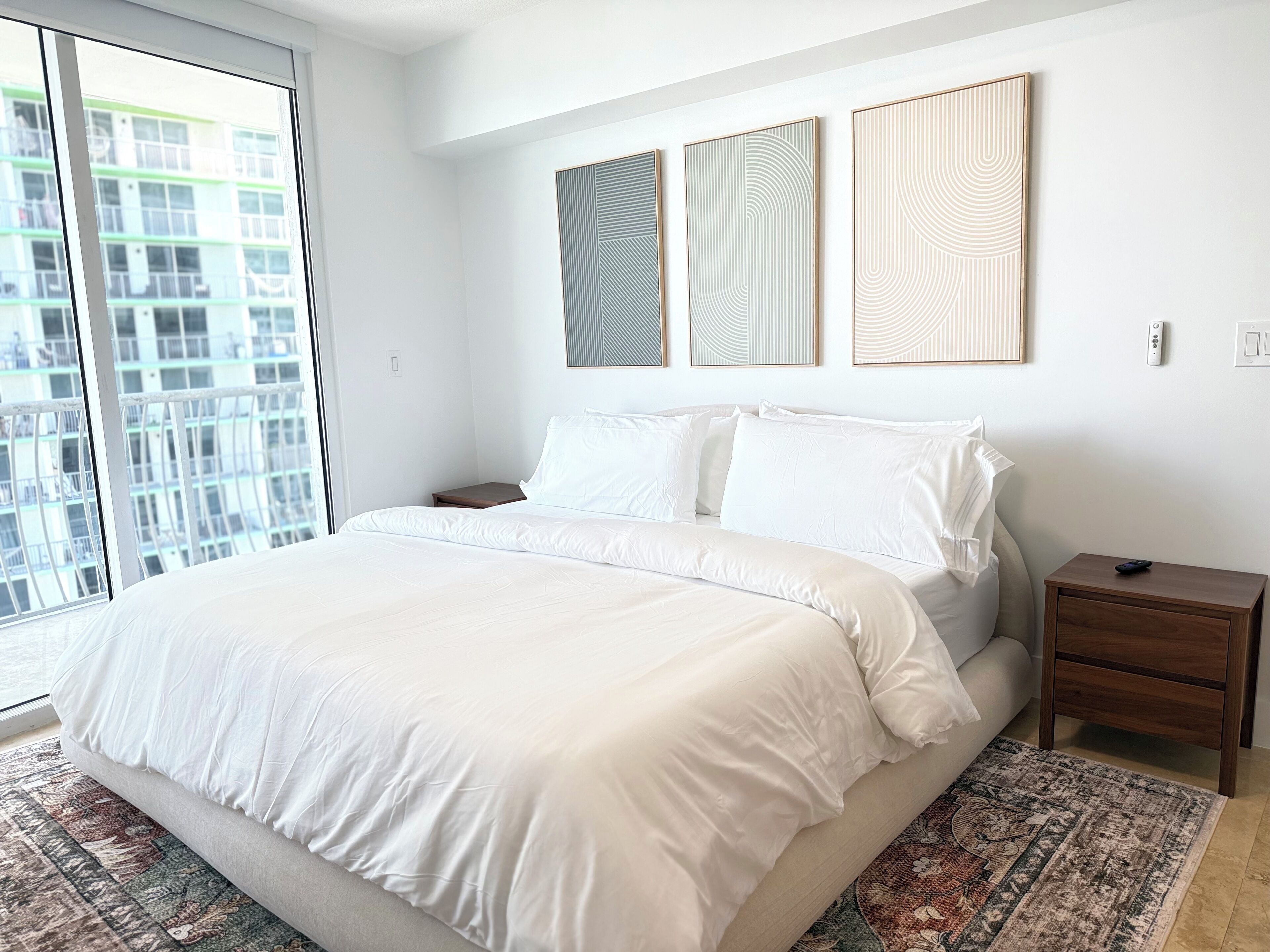 Un dormitorio con una cama grande, una mesita de noche, un panel de control en la pared y vistas de un edificio a través de una ventana.