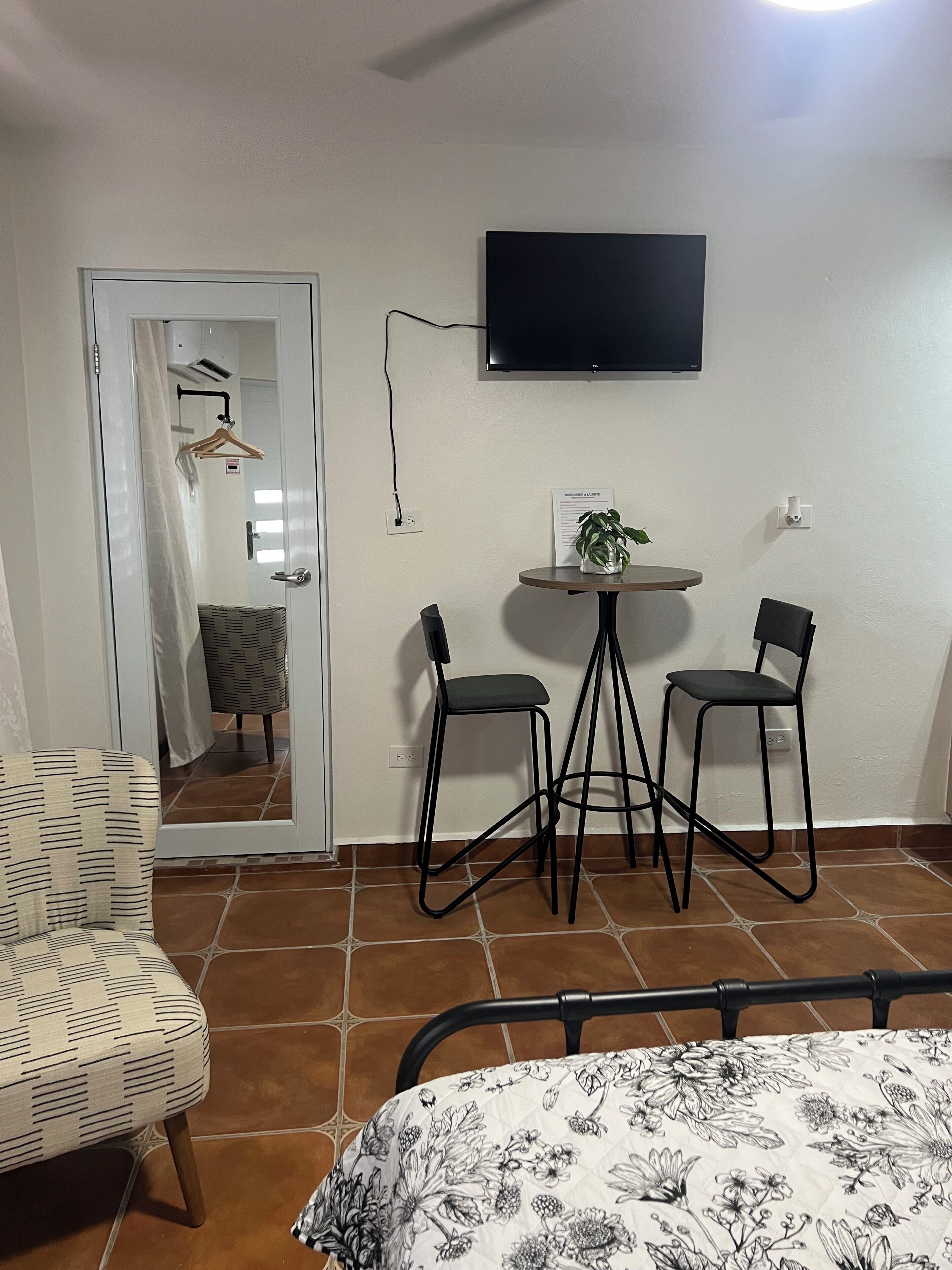 A room with a bed, a TV mounted on the wall, two chairs around a small table, and a door leading to another room.