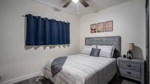 Un dormitorio con cama, mesita de noche, lámpara, ventilador de techo y ventana con cortinas.