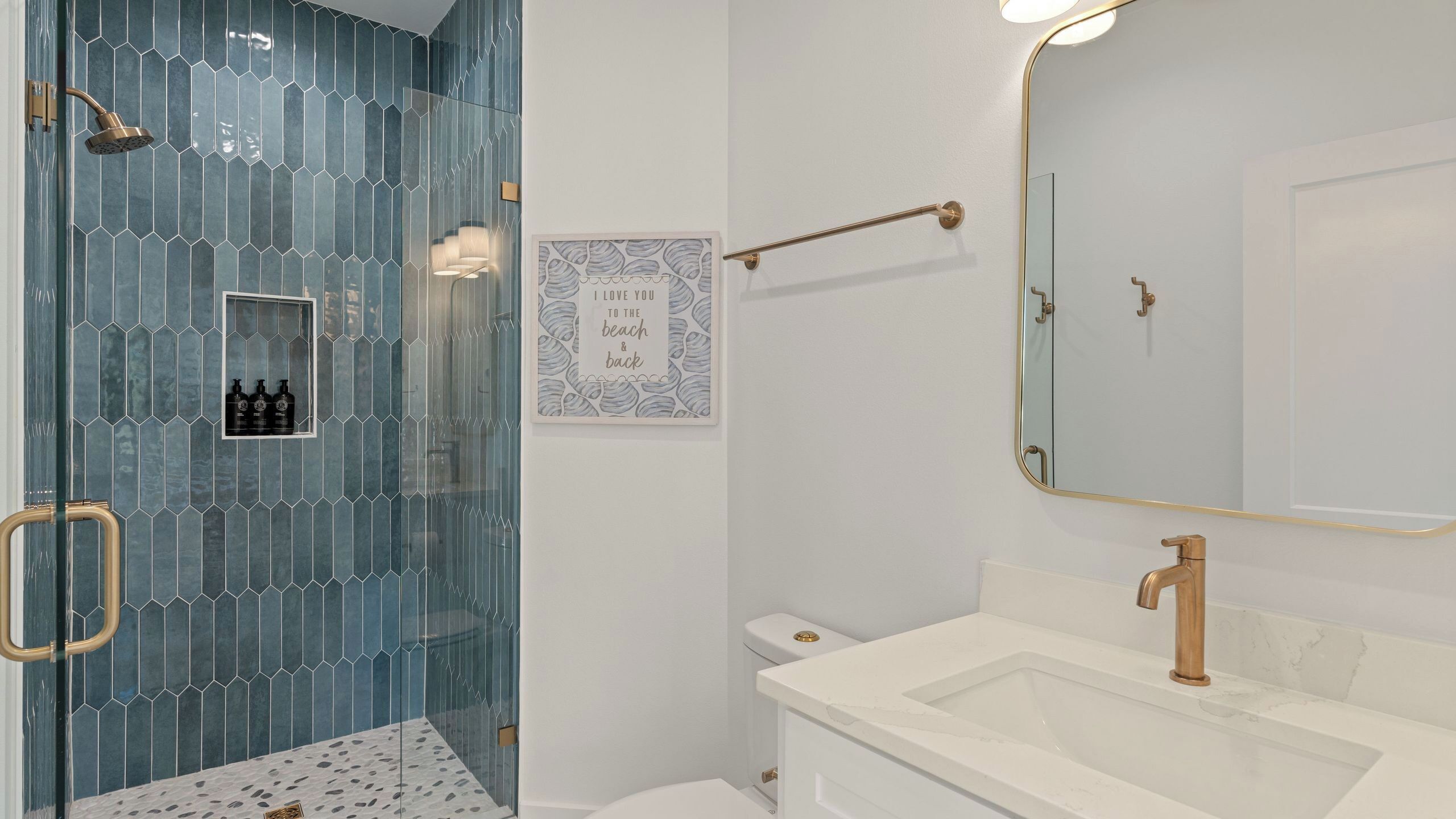Un baño con ducha acristalada, lavamanos blanco, espejo grande y una cita enmarcada en la pared.