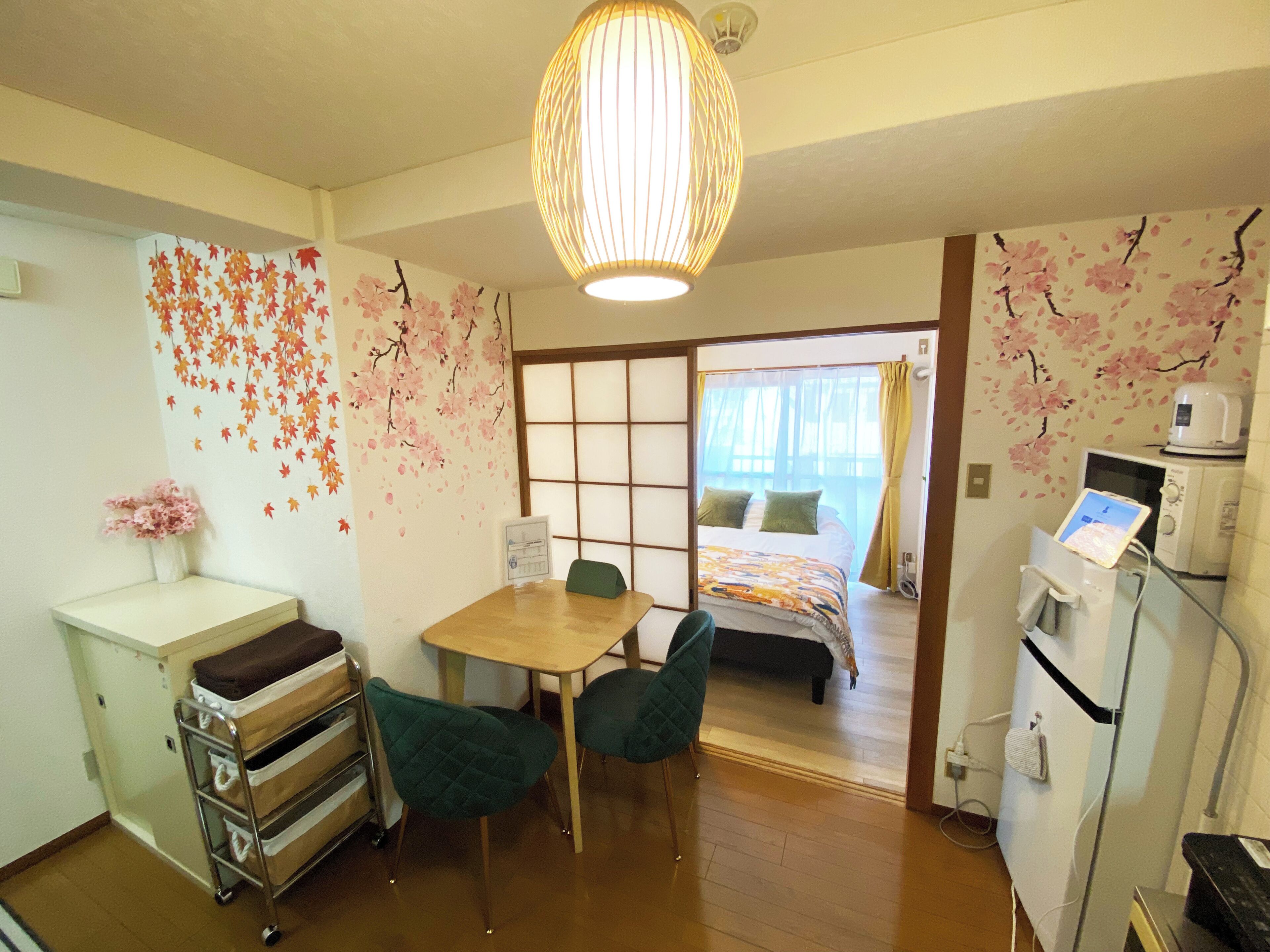 Ein Hotelzimmer mit Holztisch, zwei Stühlen, einem Bett mit bunter Bettdecke, einem Wandheizkörper, einer kleinen Kommode und einer Wand mit Kirschblütenmotiven.
