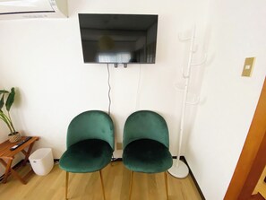 A room with a flat-screen TV mounted on the wall, two green chairs, a small wooden table with a remote control, a white trash bin, and a white floor lamp. - 1 minute walk from the station | Many supermarkets and restaurants | Atmospheric downtown area | Easy access to Shibuya/Kamata, etc. (Yaguchi Ota-ku)
