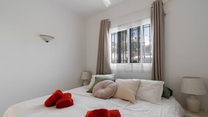 Un dormitorio con cama, dos mesitas de noche, una ventana con cortinas e iluminación en la pared.