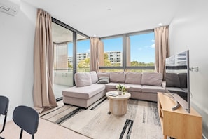 A modern living room with a sectional sofa, a coffee table, a TV, and large windows with curtains. - Modern 2BR Living with EV Charging and 7-Min to Tram (Dickson)