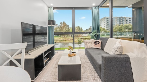 Een moderne woonkamer met een grote televisie, een bank, een salontafel en uitzicht op een park en gebouwen door grote ramen.