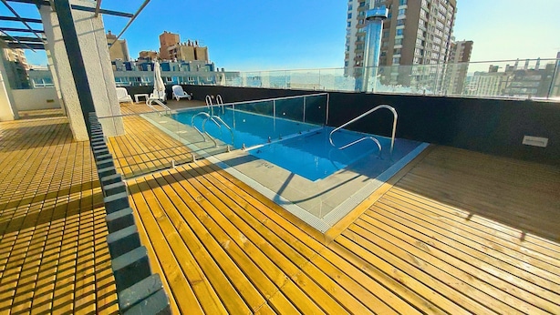 Une piscine sur le toit, dotée d’une terrasse en bois, de garde-corps en métal et offrant une vue dégagée sur le panorama urbain.