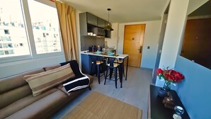 Un salon avec un canapé, un coin repas avec une table et des chaises, un espace cuisine et une vue sur les bâtiments environnants.