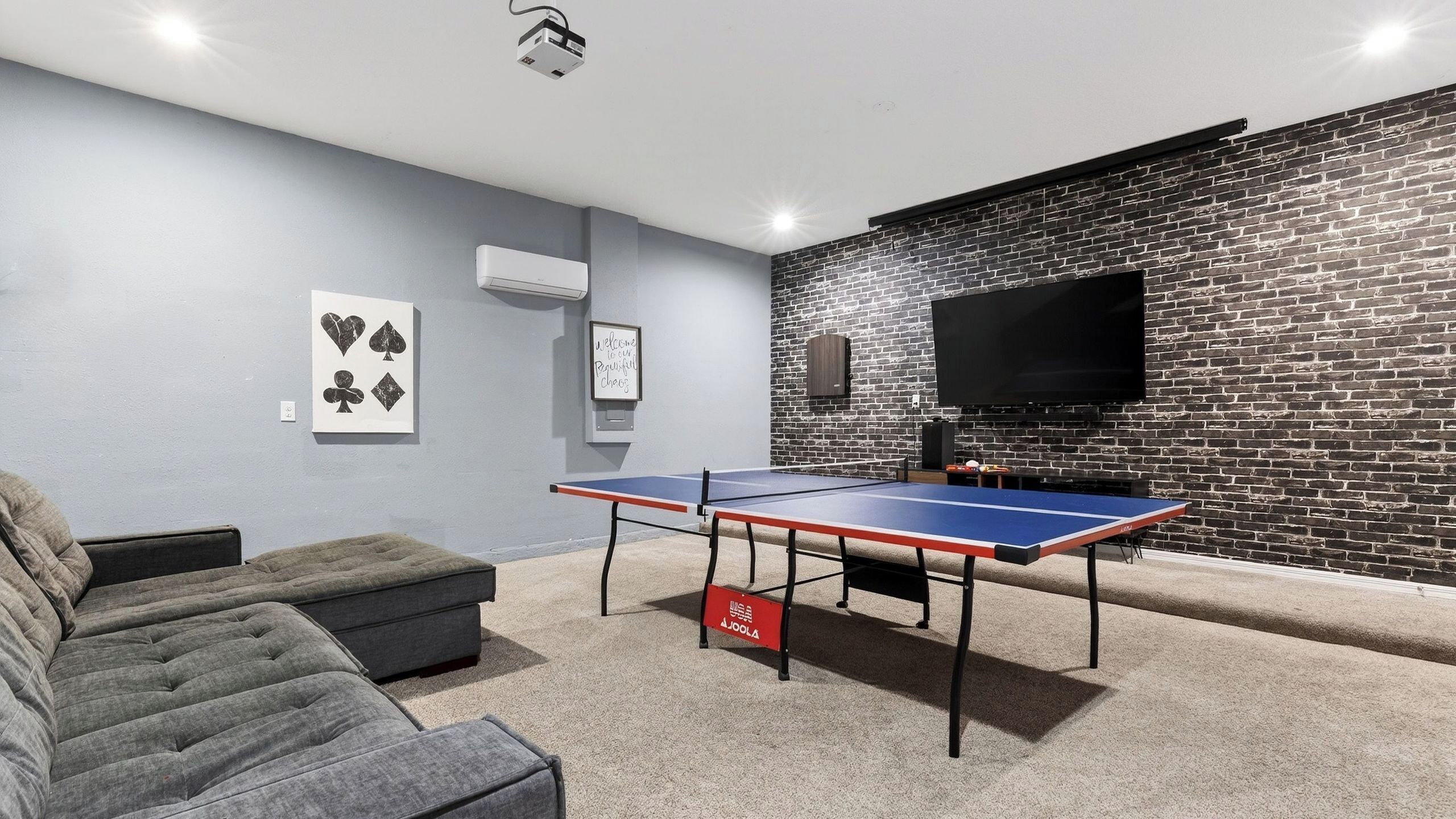 A room with a ping pong table, a flat-screen TV, a brick wall, and a gray couch.